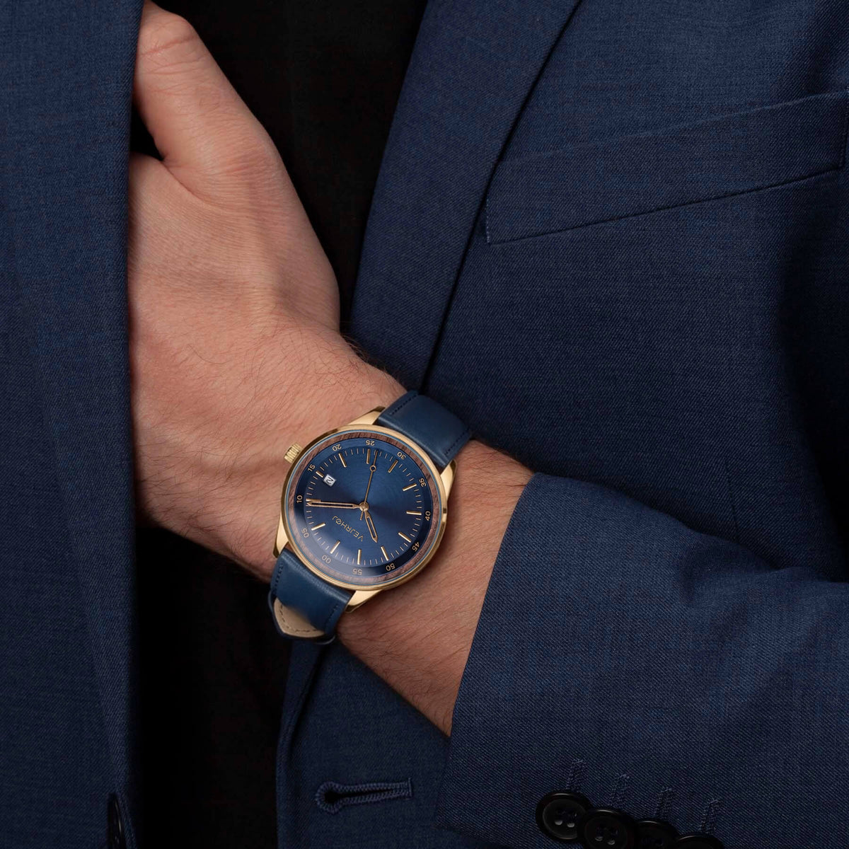 A close-up of a men's wristwatch with a blue dial and blue strap, worn on a person's wrist. The watch has a gold-tone case and is styled on a person wearing a dark blue suit jacket and a black shirt.