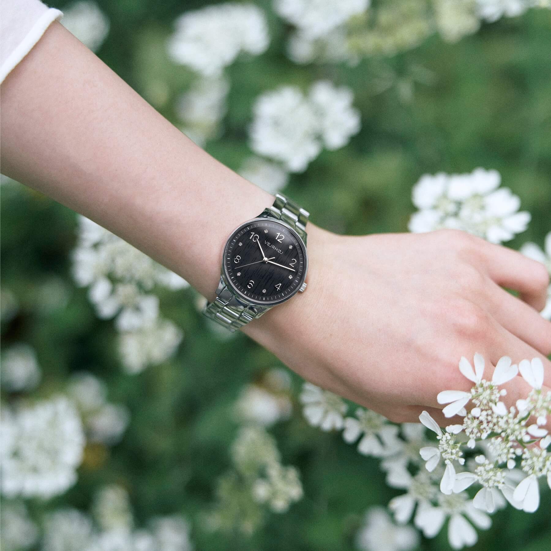 
VEJRHØJ's Petite Royal Black, a ladies' watch with a black wood grain face that looks like it blends in with the grass and flowers in natural light